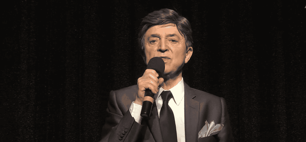 Prezentacja na scenie, mężczyzna w eleganckim smokingu, trzyma mikrofon, występ na koncertach i wydarzeniach kulturalnych.