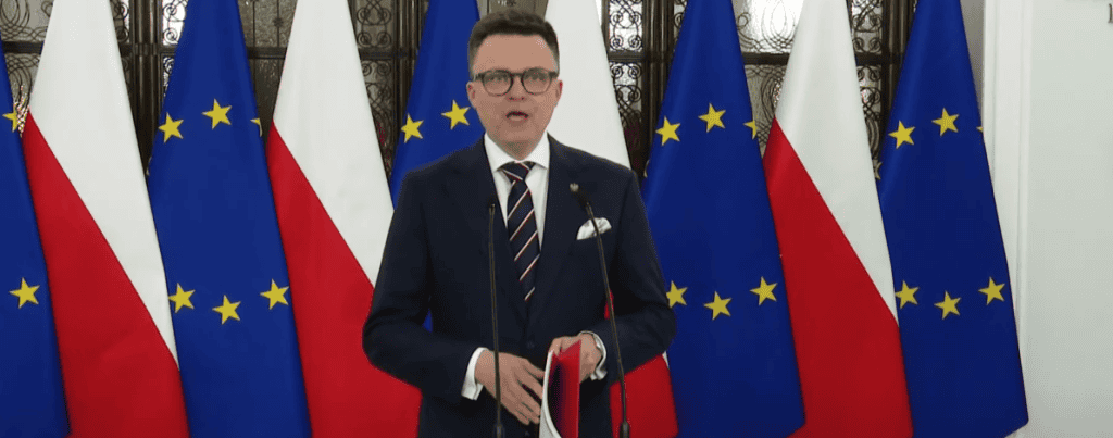 Flaga polskiej i unijnej, polityk podczas przemówienia, flaga na tle, oficjalne wydarzenie, patriotyczne tło.