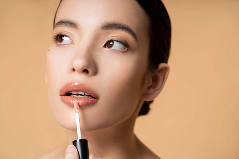 Naturalny makijaż ust z błyszczykiem, kobieta w bluzce w neutralnych kolorach, pastelowe tło, beauty i pielęgnacja, delikatne ustylizowane, elegancki look, makijaż dla codziennego użytku, profesjonalna aplikacja kosmetyków, idealny na co dzień i na wyjątkowe okazje.